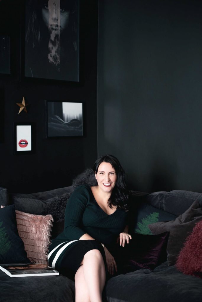 Large portrait photo of the CEO of D&R, Ally Dowsing-Reynolds, sitting on a dark grey sofa in her home.