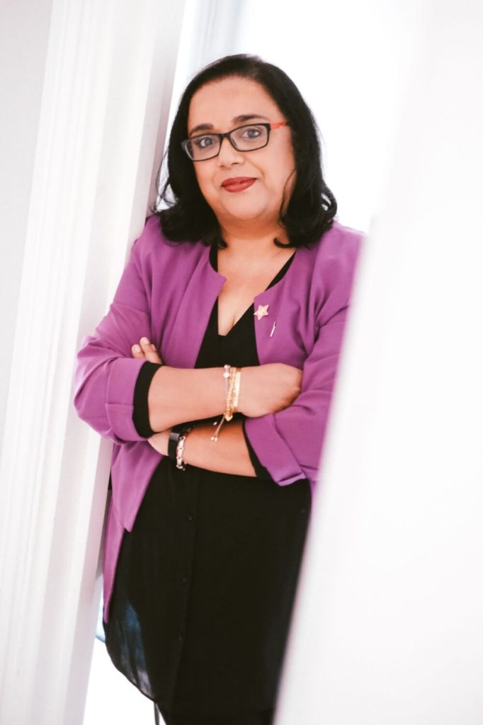 Advita Patel wearing a purple jacket and leaning in the doorway of a white room.